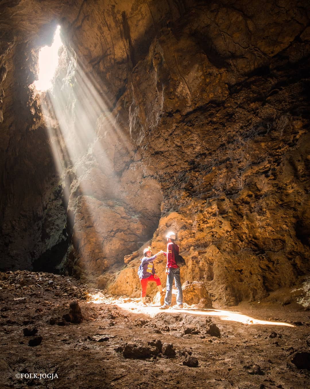 Two people in helmets stand inside Cokro Cave, bathed in sunlight streaming through the vertical shaft.