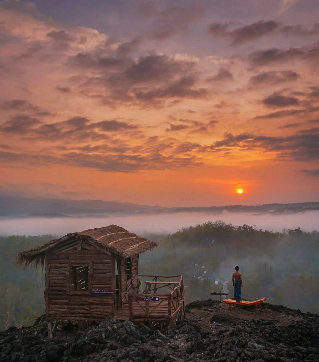 A person standing near a wooden house on Ireng Mountain, watching the sunrise over a misty landscape.