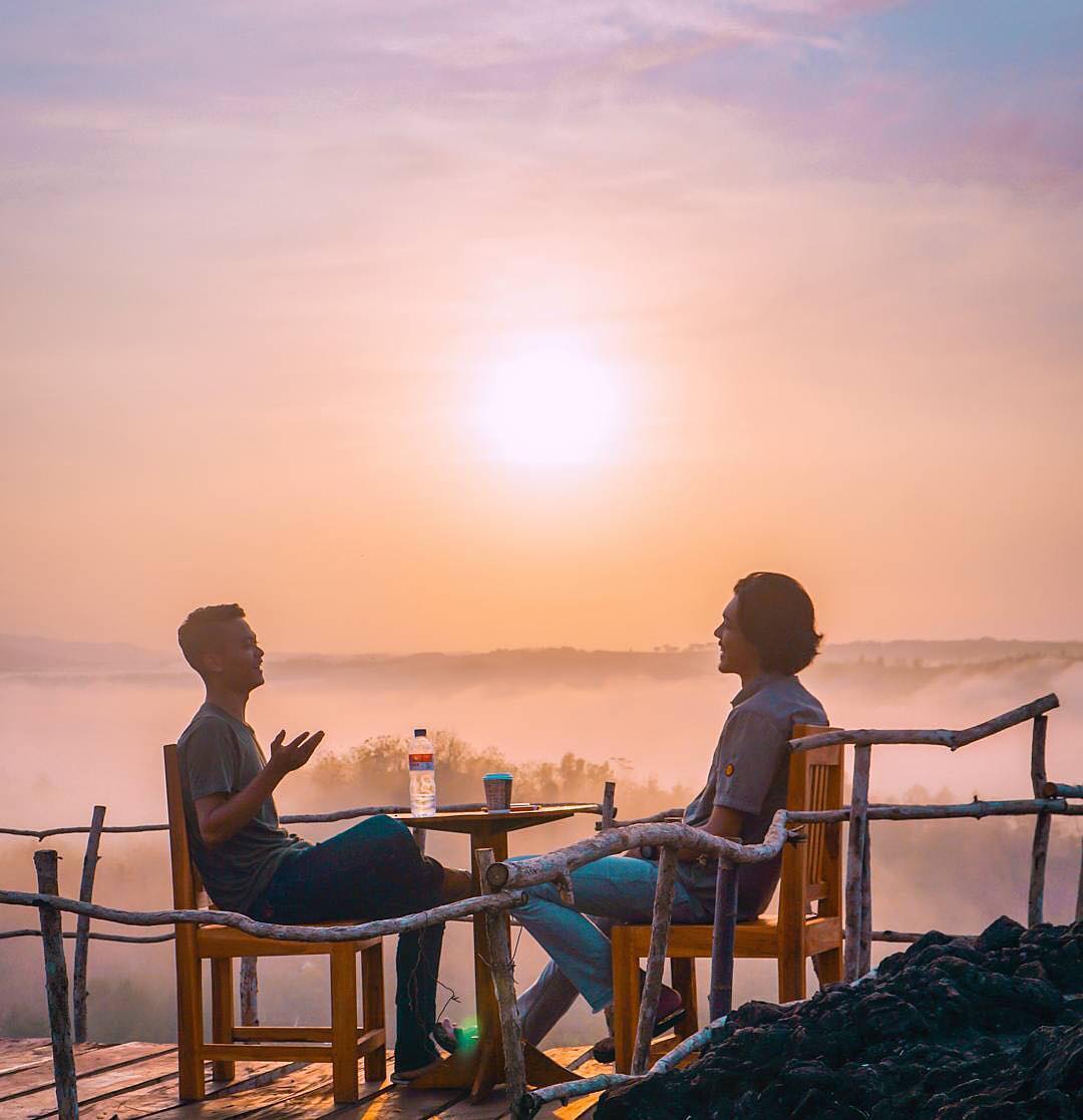 Two people sitting at a wooden table on Ireng Mountain, enjoying a conversation during a golden sunrise with a misty landscape in the background.