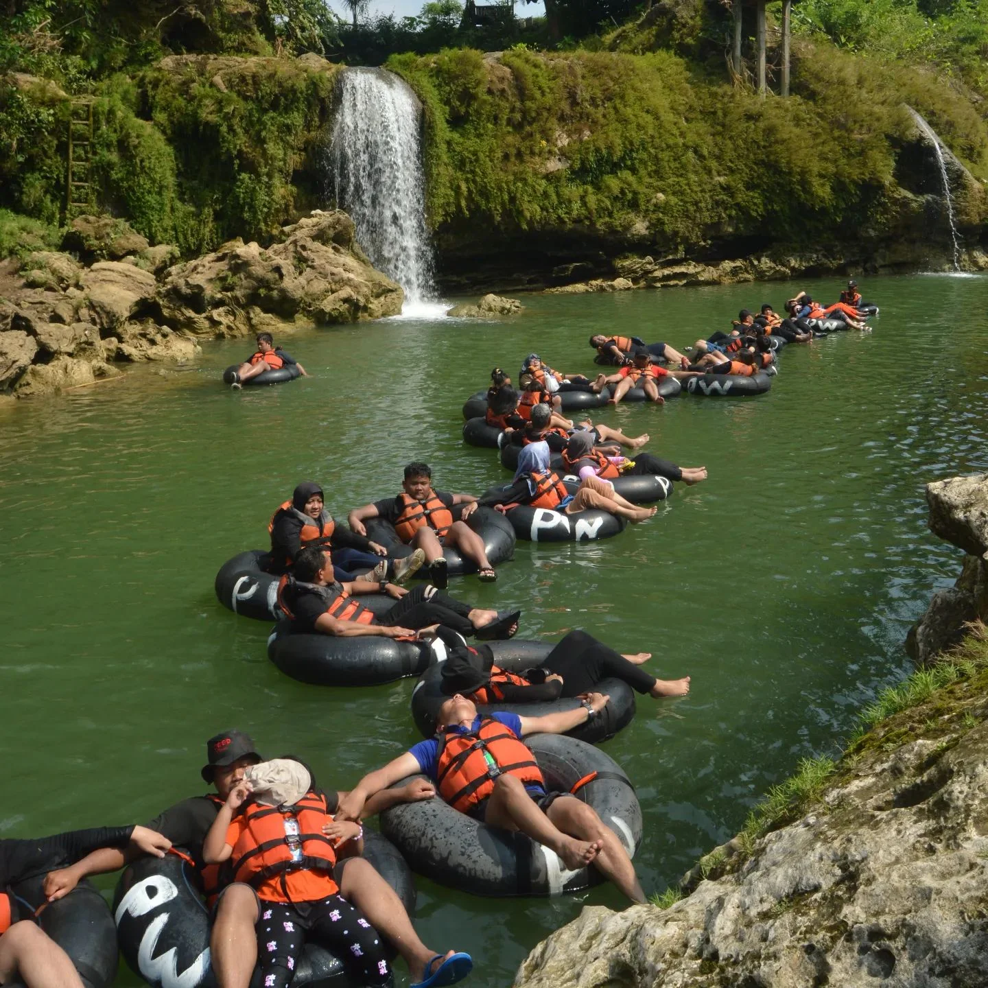 Visitors floating in a line of rubber tubes in the emerald waters in front of a waterfall at Pindul Cave.