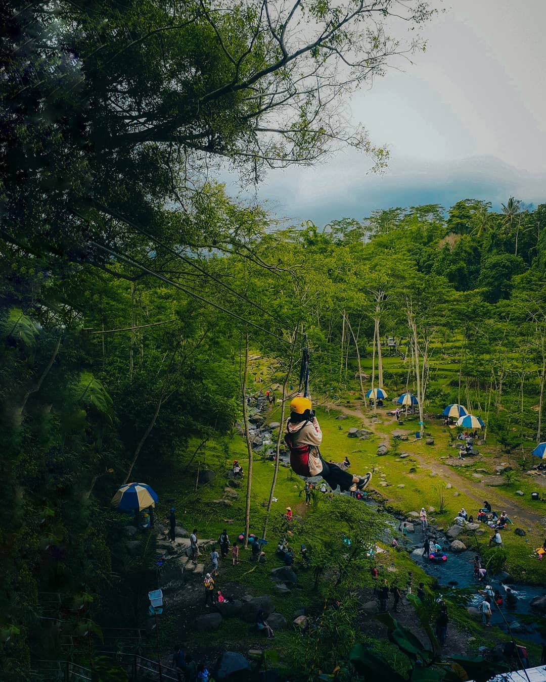 Person enjoying a zipline ride over a green park at Ledok Sambi, with families below.