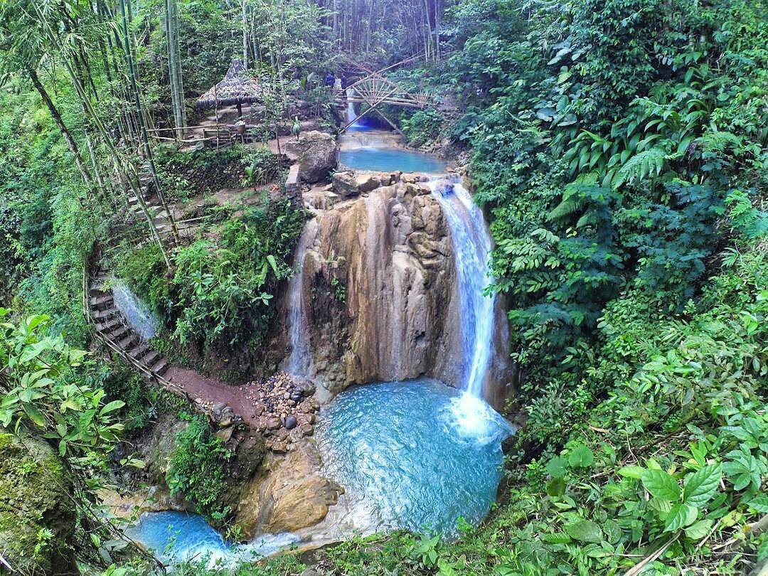 A panoramic view of Kedung Pedut Waterfall showcasing multiple levels of turquoise pools, bamboo structures, and lush green vegetation.