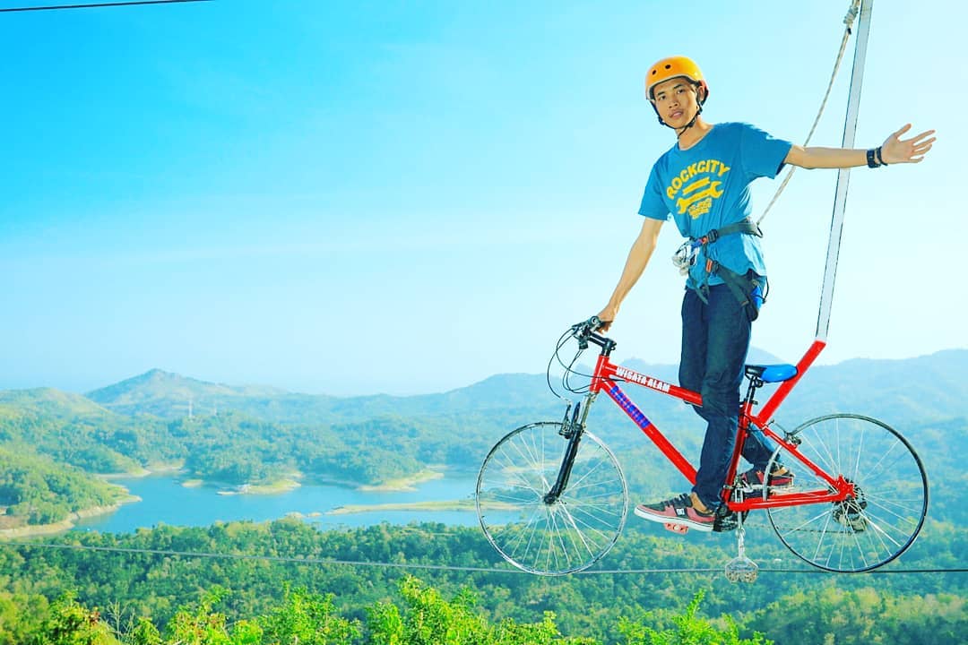 A person riding a bicycle on a high wire with safety harnesses, overlooking the lush green hills and Sermo Reservoir at Kalibiru Natural Tourism in Yogyakarta, Indonesia.