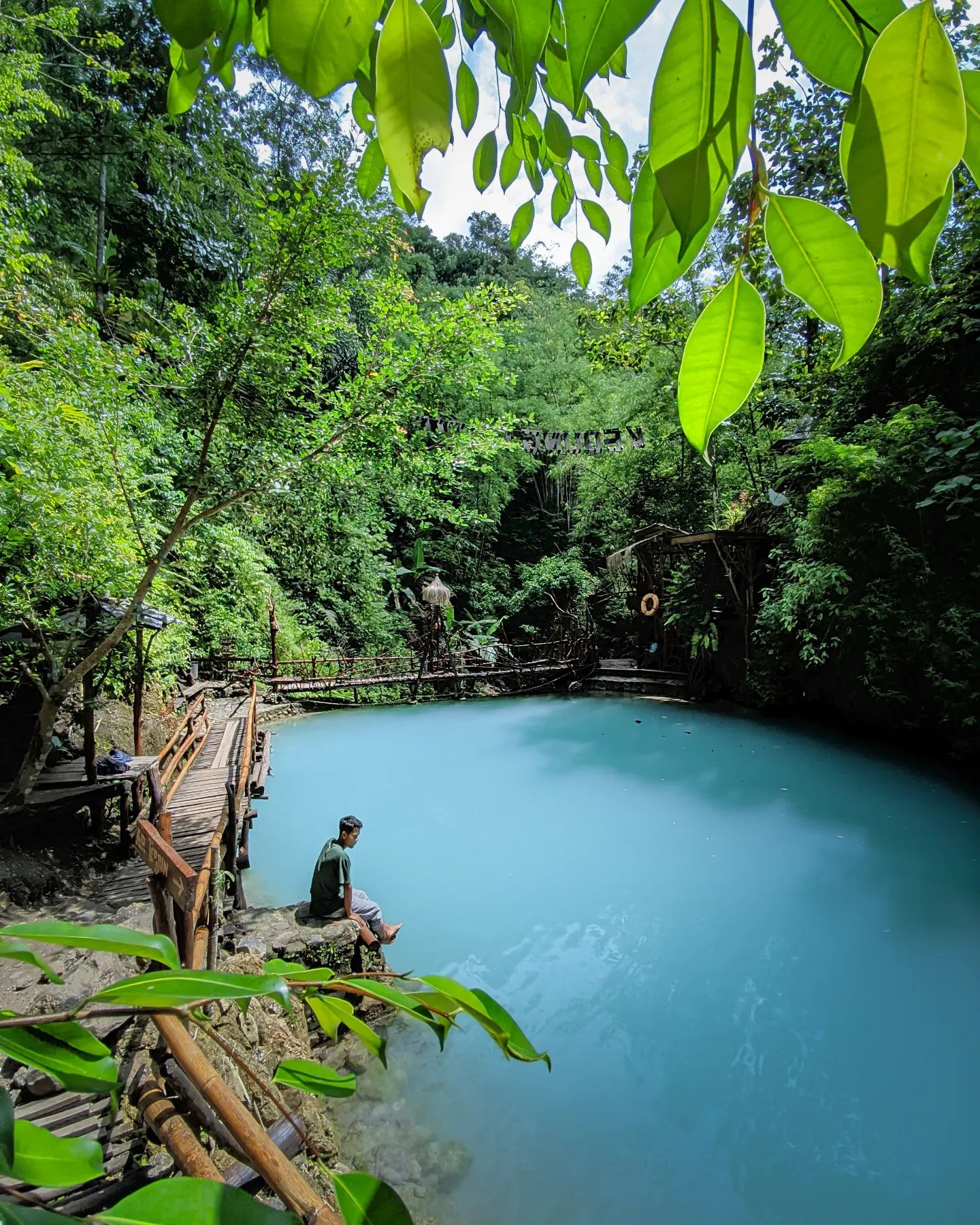 A person sitting by the edge of a calm, turquoise pond surrounded by lush greenery and wooden walkways at Kedung Pedut Waterfall.