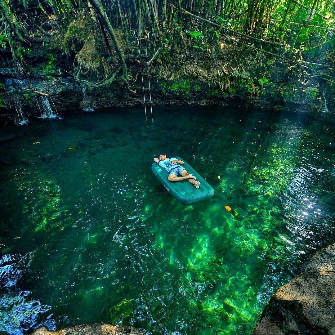 A person lounging on an inflatable float in the clear, blue-green waters of Tirta Budi Bathing Place, surrounded by lush greenery and rocky formations.