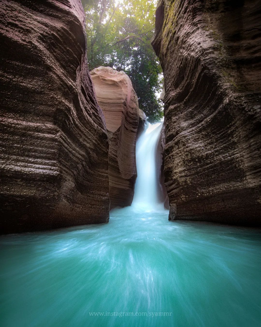 Luweng Sampang Waterfall with its clear, blue water flowing through intricate limestone cliffs surrounded by trees in Gunungkidul, Yogyakarta, Indonesia.