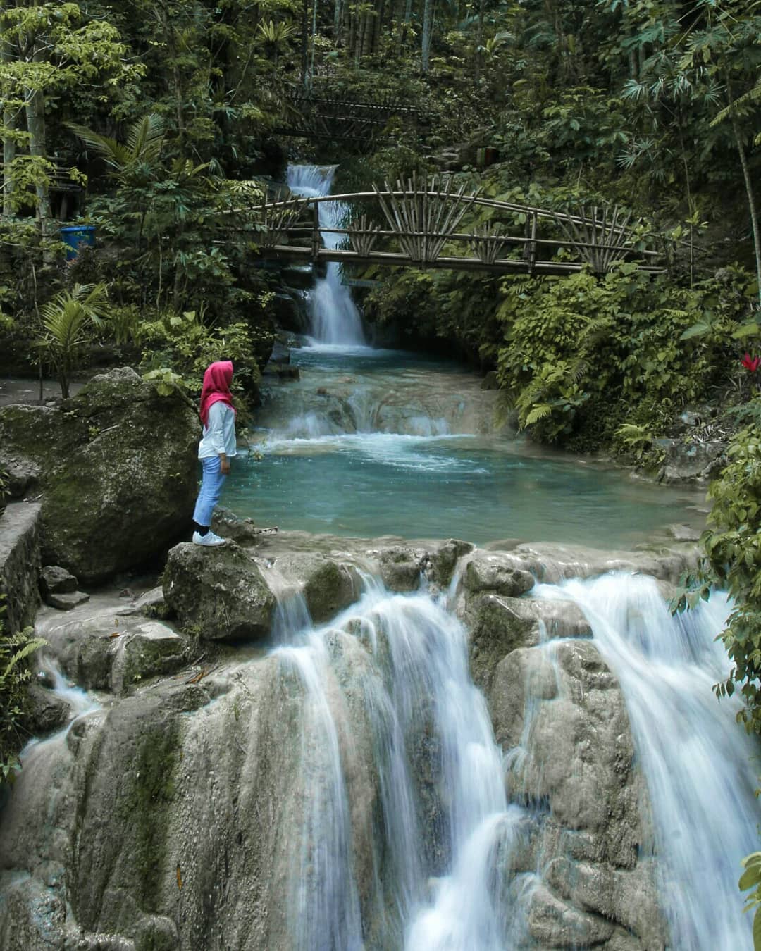 A person standing on a rock near the cascading waters of Kedung Pedut Waterfall, surrounded by lush vegetation and wooden bridges.