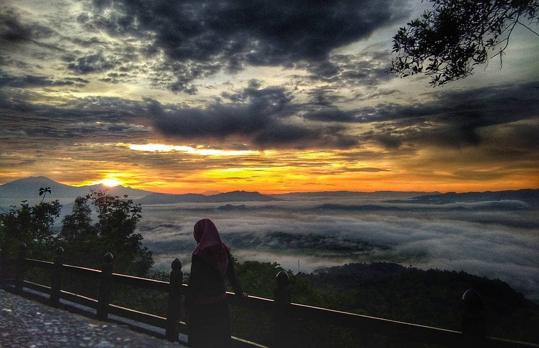 A person stands on a viewpoint at Gambar Mountain during a stunning sunrise, with clouds below and mountains in the distance.