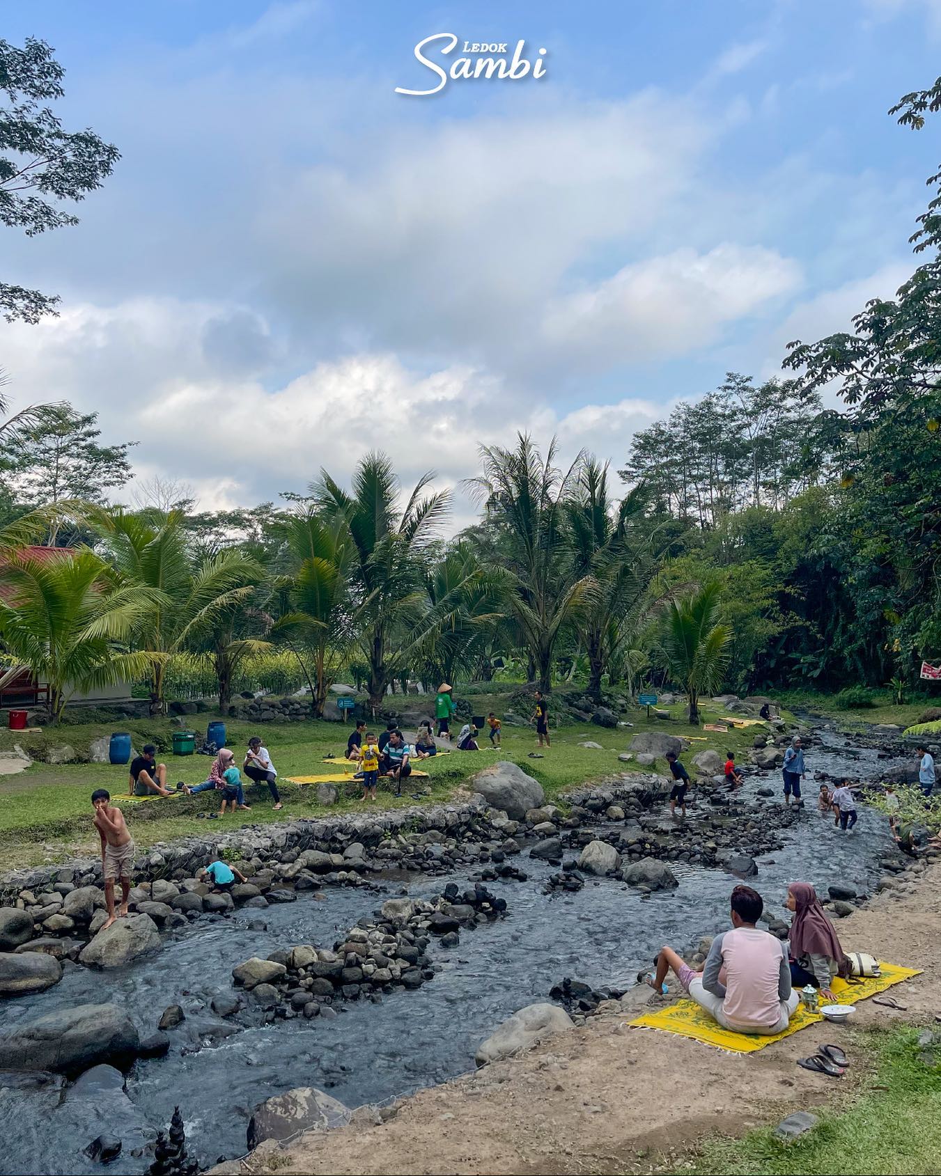 Families relaxing and playing by a small river at Ledok Sambi, surrounded by lush vegetation.