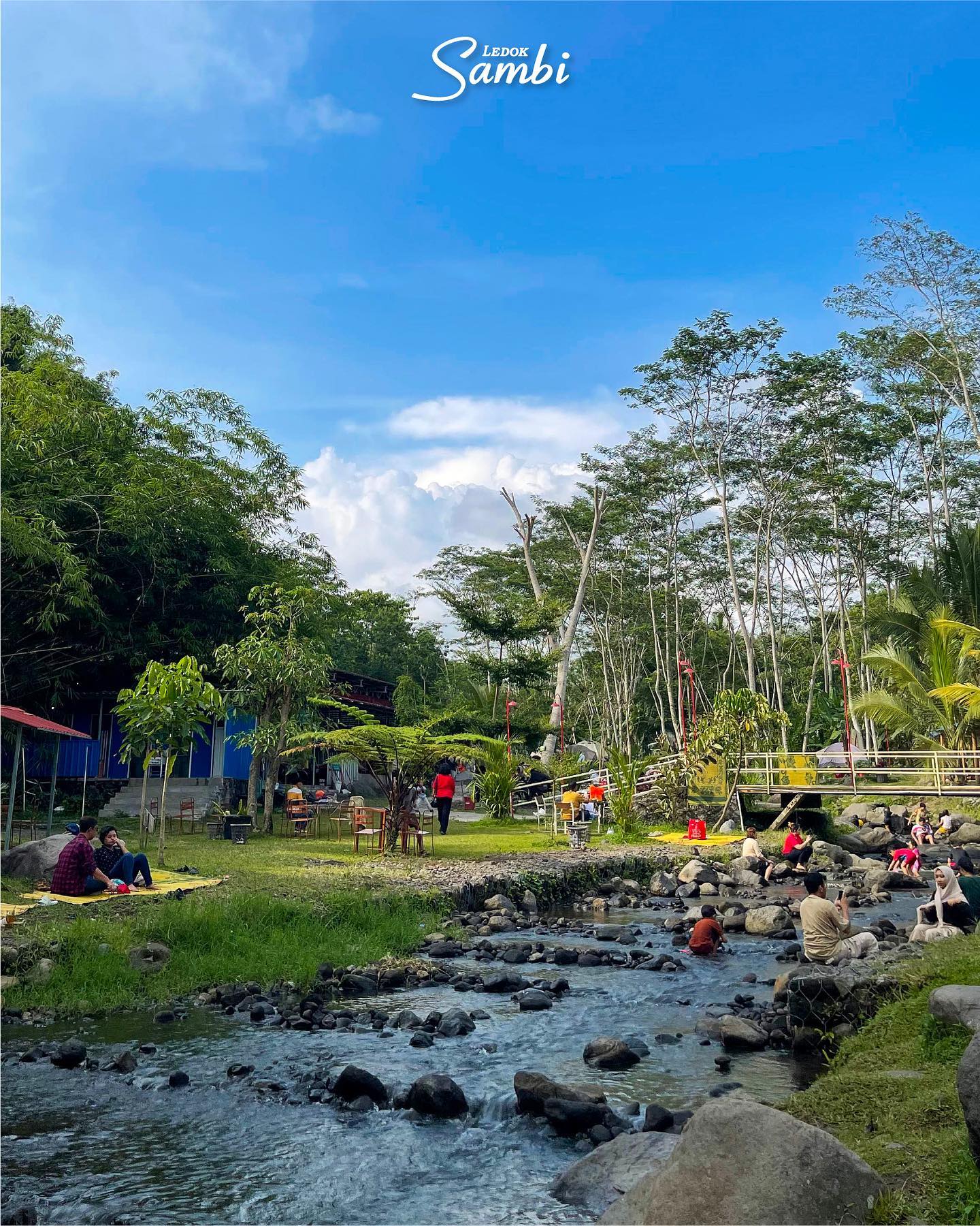 People relaxing and playing near a small river at Ledok Sambi, surrounded by green trees and grass under a bright blue sky.