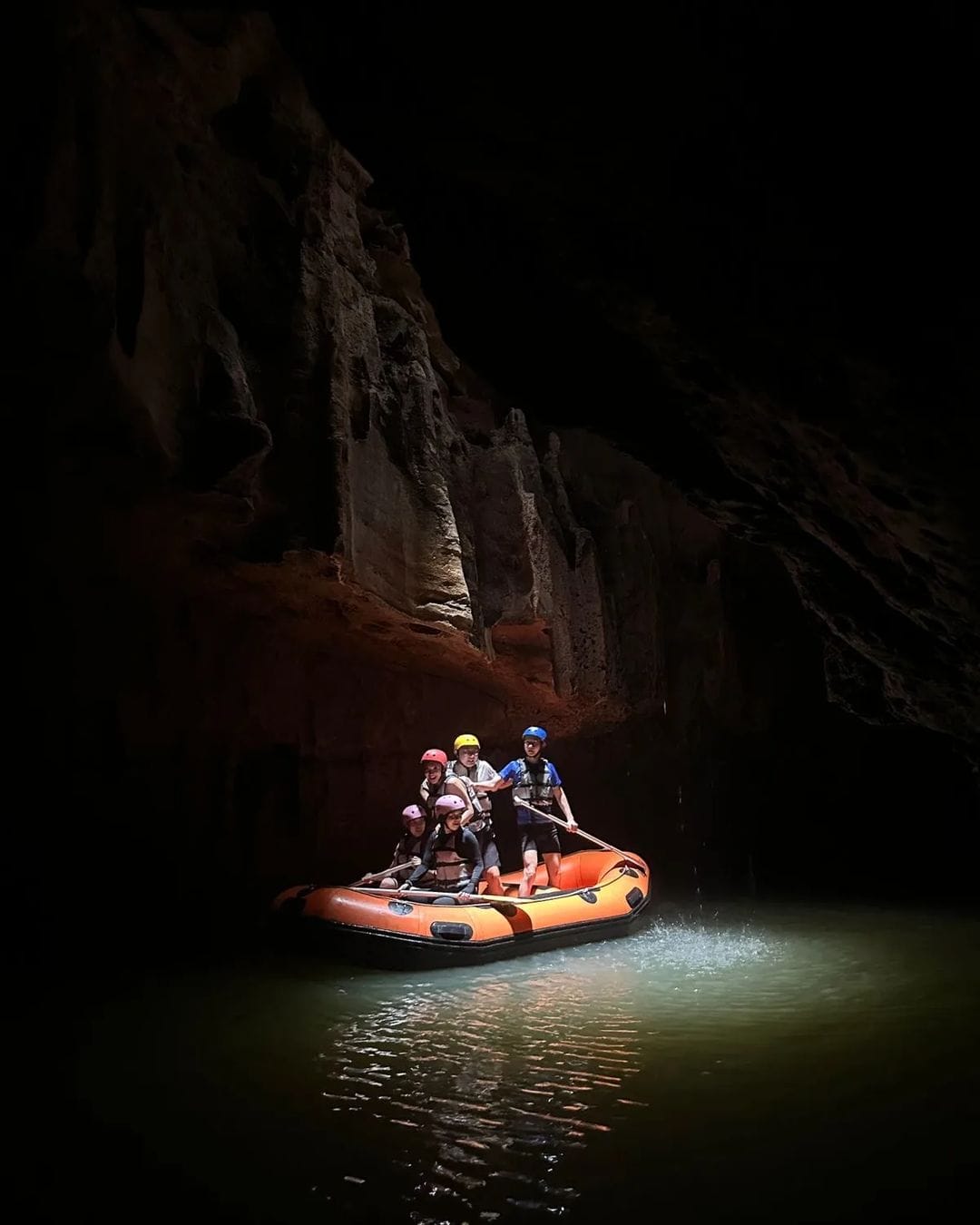 A group of tourists in an orange inflatable raft paddle through the underground river in Tanding Cave, surrounded by dark cave walls illuminated by dim light.