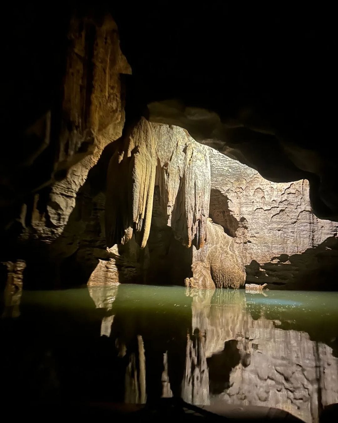 Dim light reveals reflections of stalactites and rock formations in the still underground river inside Tanding Cave.