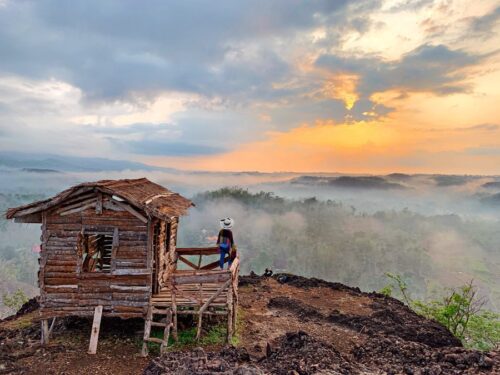 A person standing near a wooden house on top of Ireng Mountain, enjoying the golden sunrise over a misty landscape.