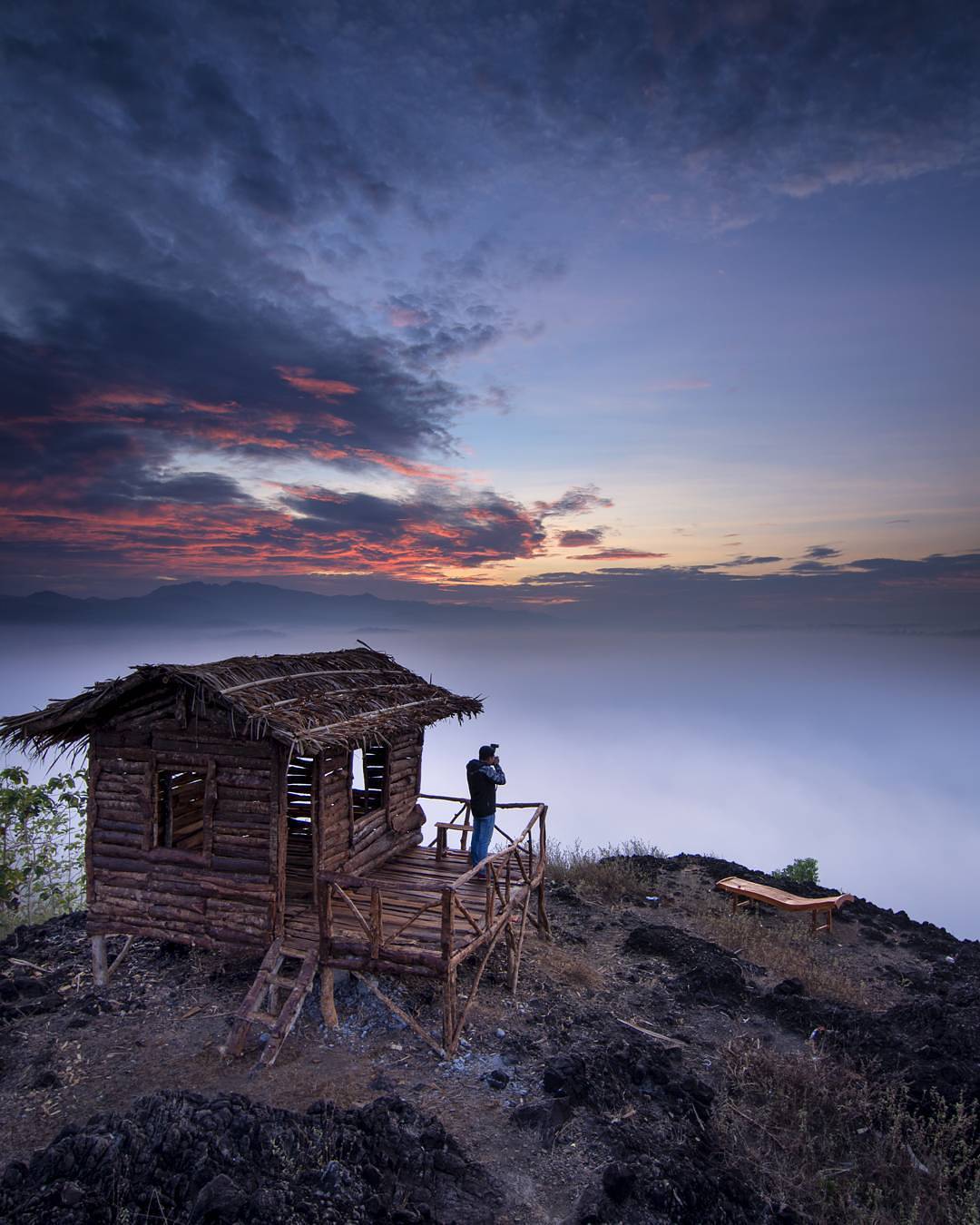 A person taking photos from the porch of a wooden house on Ireng Mountain during dawn, with a dramatic sky and misty landscape below.