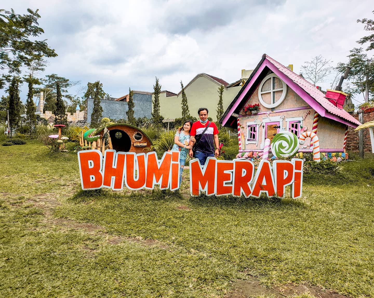 Two visitors standing beside a colorful sign that reads "Bhumi Merapi" in front of a whimsical house at Bhumi Merapi Agrotourism.