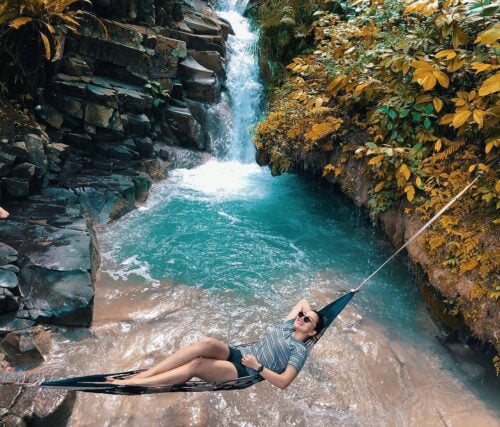 A person relaxing in a hammock near the blue-green pond and waterfall at Kedung Pedut Waterfall.