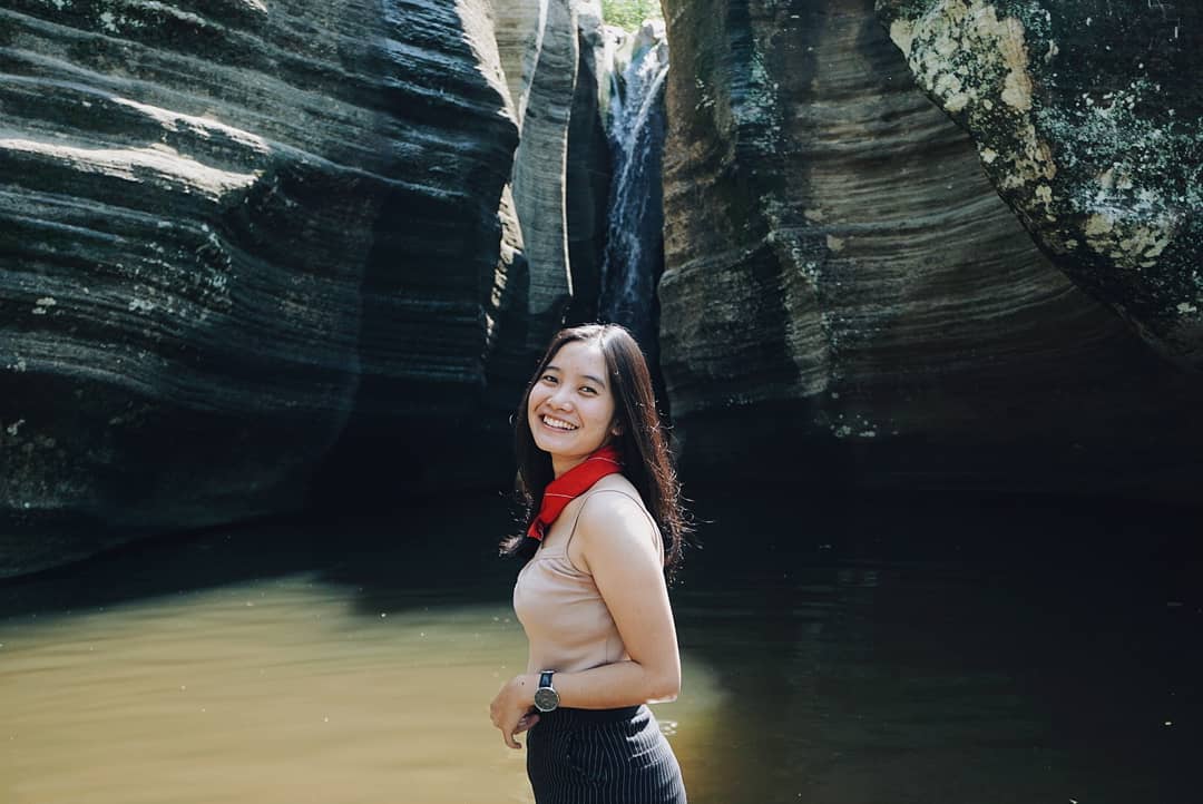 A young woman smiling and standing in front of Luweng Sampang Waterfall with unique limestone cliffs in Gunungkidul, Yogyakarta, Indonesia.