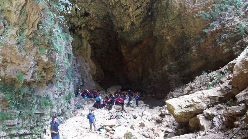 A group of people explore the entrance of Ngeleng Cave, surrounded by rough rock formations and lush greenery.