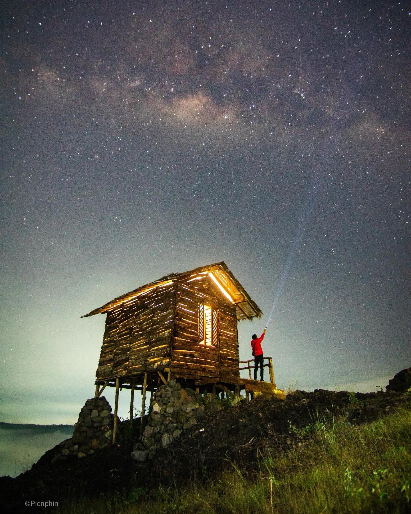 A person standing on the porch of a wooden house on Ireng Mountain, shining a flashlight towards the starry night sky.