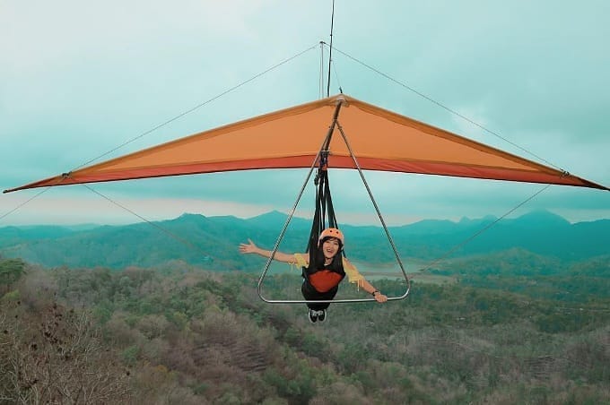 A person enjoying a hang gliding experience with safety gear, soaring over the lush green hills at Kalibiru Natural Tourism in Yogyakarta, Indonesia.