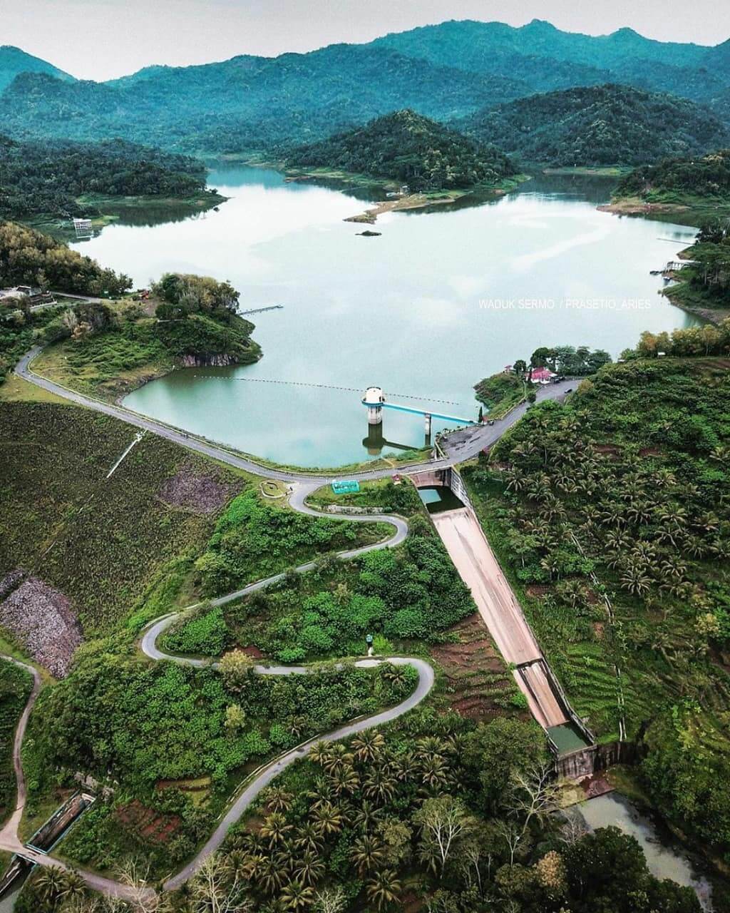 Aerial view of Sermo Reservoir surrounded by lush green hills and winding roads.