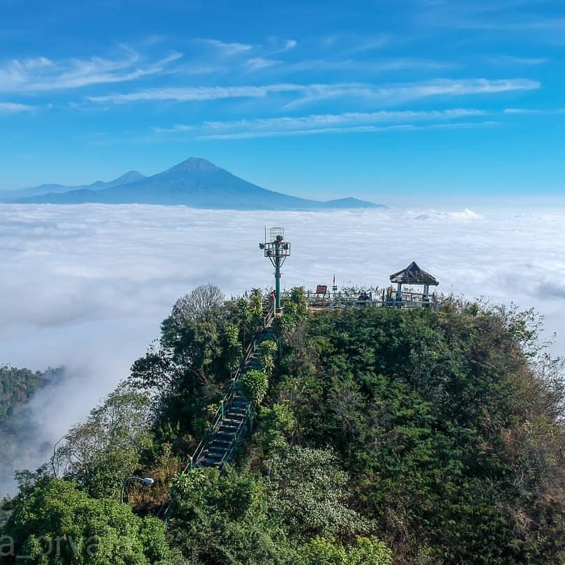 A scenic view from Suroloyo Peak, Yogyakarta, Indonesia, showing a pavilion atop the peak with fluffy clouds below and Mount Merapi and Mount Merbabu in the background.