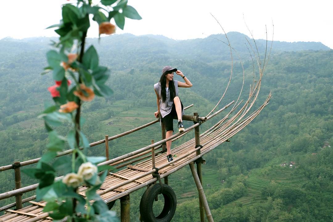 A woman standing on a bamboo viewing platform with lush green hills in the background at Mojo Gumelem Hill in Yogyakarta, Indonesia.