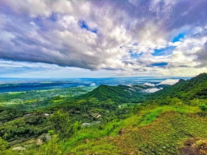 A panoramic view from Gunung Gambar showcasing lush green hills, expansive forests, and a partly cloudy sky.