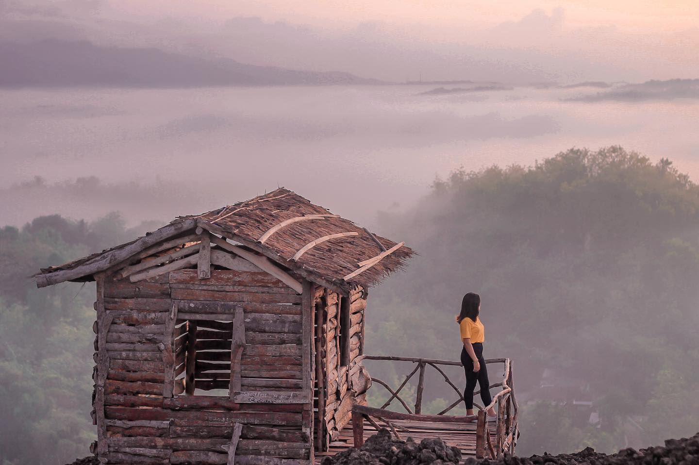 A woman standing on the porch of a wooden house on Ireng Mountain, overlooking a misty landscape during sunrise.