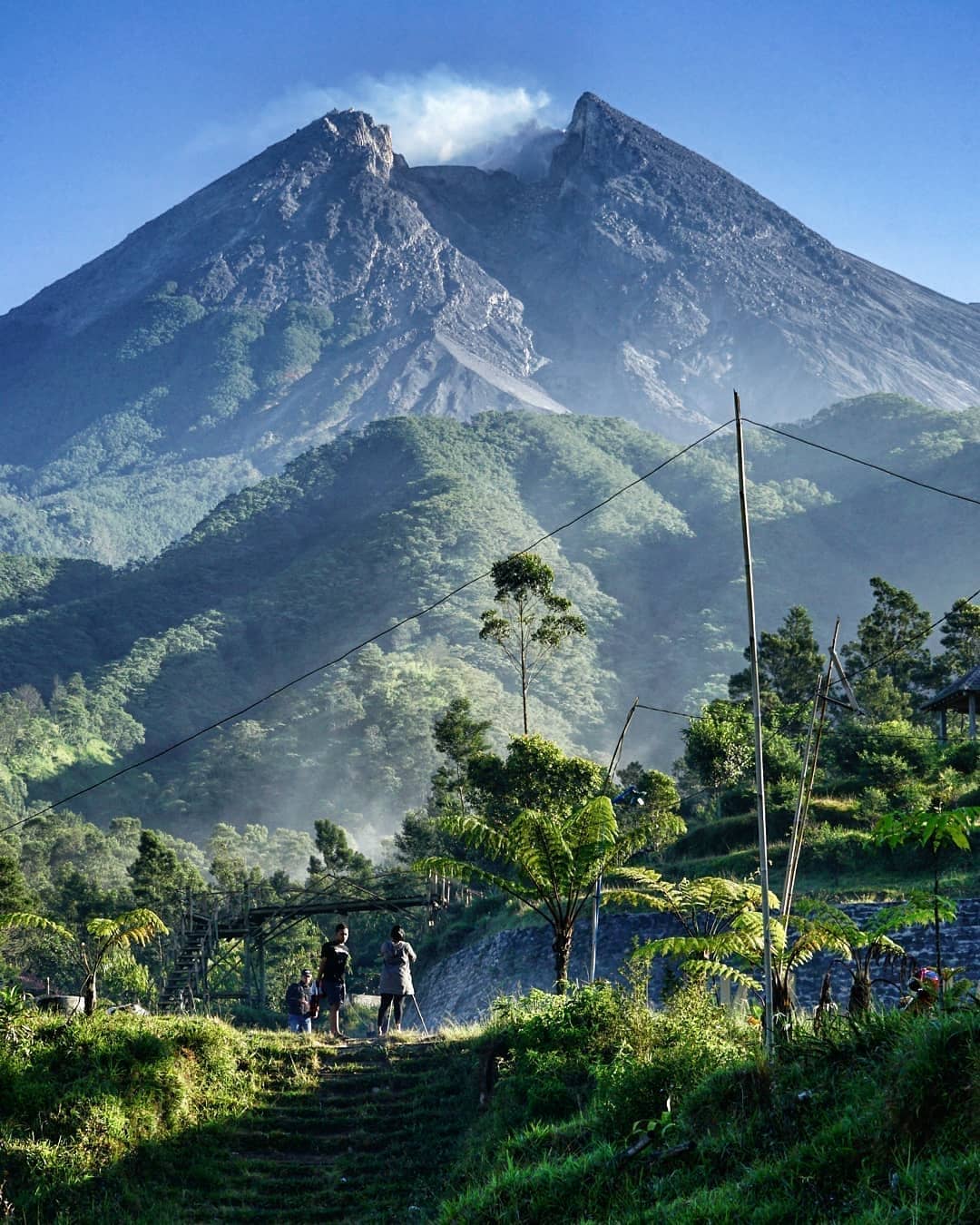 People standing on a grassy path at Klangon Hill with Mount Merapi towering in the background under a clear blue sky.