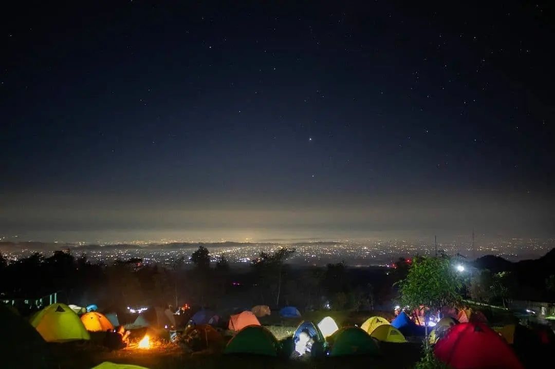 Campers in colorful tents at Klangon Hill, enjoying a campfire under a starry night sky with city lights in the distance.