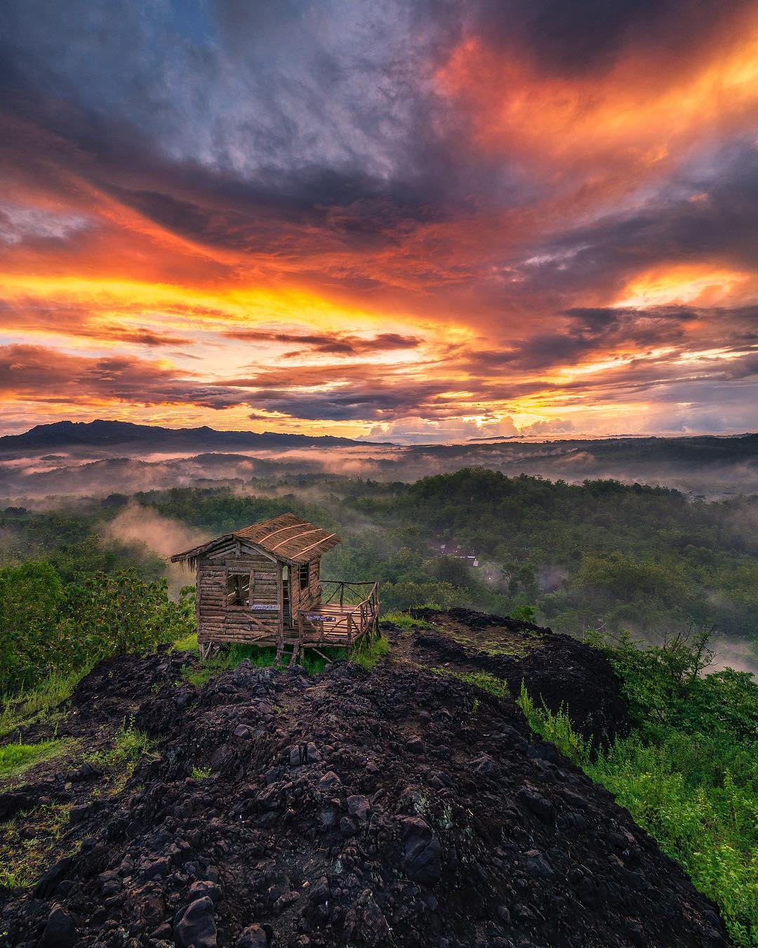 A wooden house on the dark volcanic rocks of Ireng Mountain, illuminated by the vibrant colors of the sunrise, with misty hills in the background.