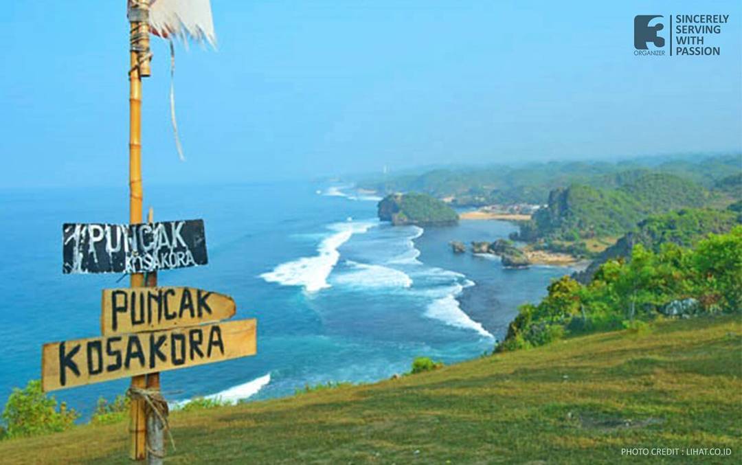 A wooden signpost on Kosakora Peak pointing to "Puncak Kosakora" with a beautiful coastal landscape in the background.