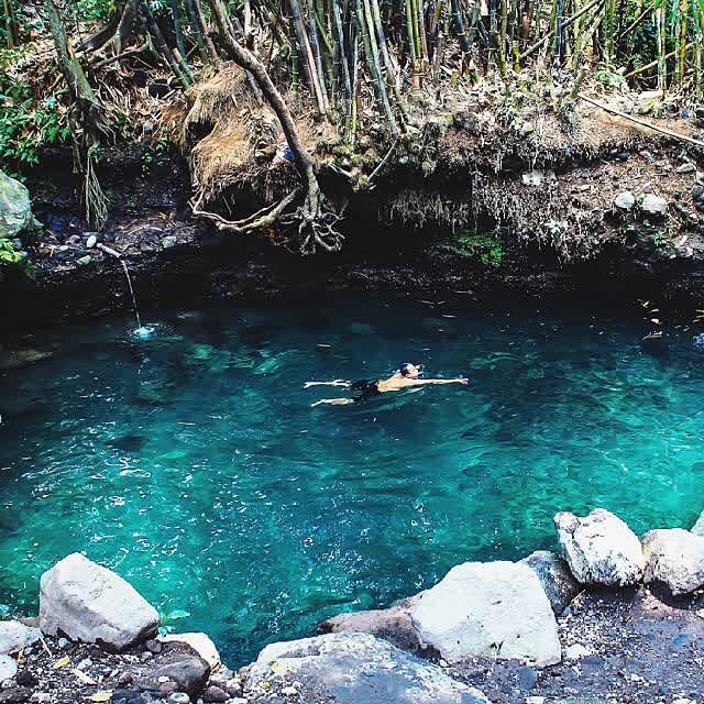 A person floating on their back in the clear blue waters of Tirta Budi Bathing Place, surrounded by rocks and dense vegetation.