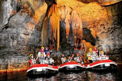 A group of tourists wearing safety gear and helmets enjoy cave rafting inside Tanding Cave, surrounded by majestic rock formations and stalactites.