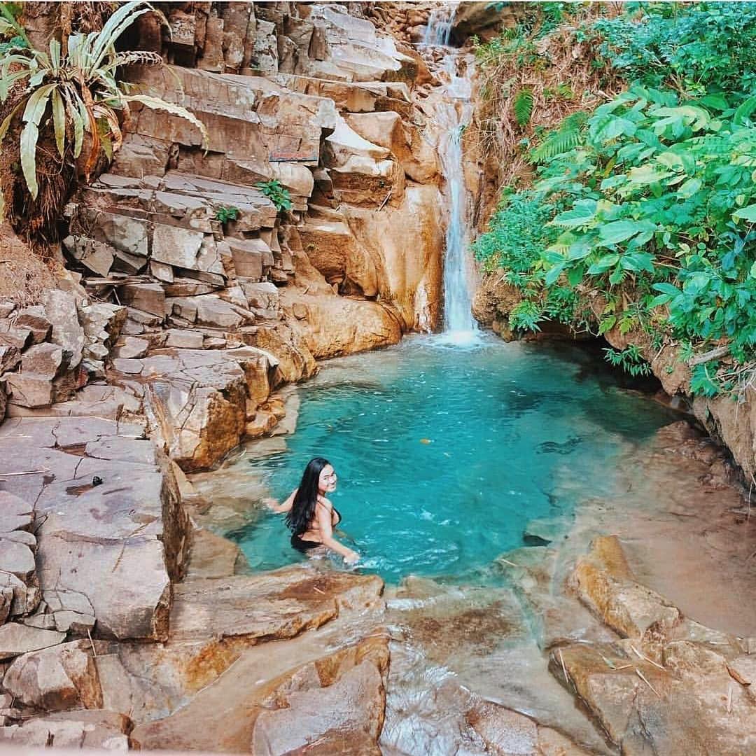 A person swimming in a crystal-clear blue-green pool at the base of Kedung Pedut Waterfall surrounded by rocky cliffs and lush vegetation.