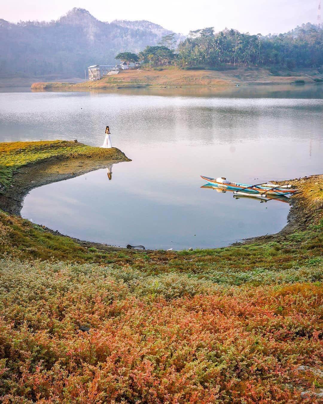 A woman in a white dress stands on the grassy shore of Sermo Reservoir with colorful vegetation in the foreground and calm water and mountains in the background.