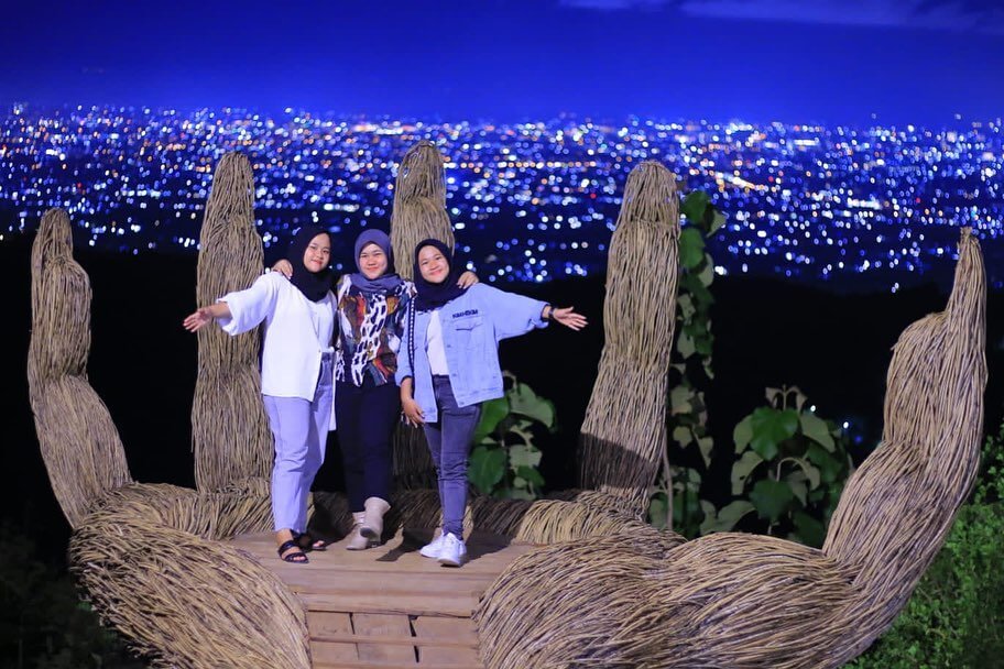 Three friends posing on the Giant Hand structure at Hutan Pinus Pengger with sparkling city lights in the background.