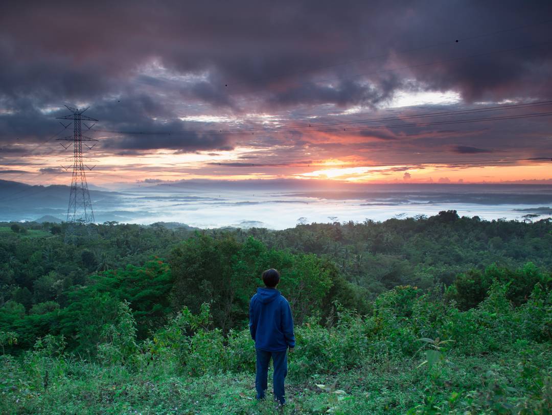 A person in a blue hoodie standing on a hill at Mount Mungker, watching a vibrant sunrise.