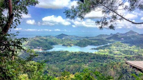 A panoramic view of lush green hills surrounding the serene Sermo Reservoir at Kalibiru Natural Tourism in Yogyakarta, Indonesia.