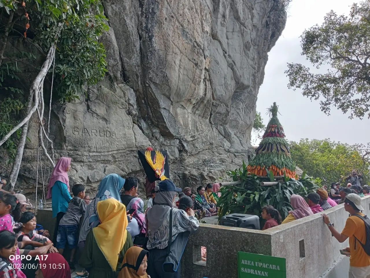 A group of people participates in the Sadranan ceremony at Gunung Gambar, with a large traditional cone-shaped arrangement of produce placed in front of a rocky cliff.