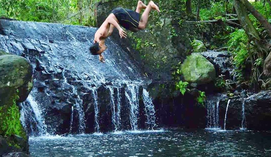 A person performing a backflip into the water from a waterfall at Tirta Budi Bathing Place, surrounded by green vegetation and rocky formations.