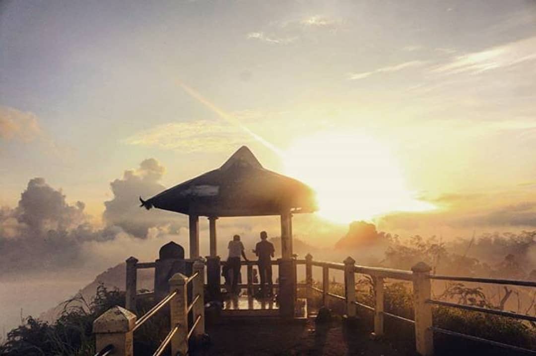 Two people enjoy a breathtaking sunrise from a pavilion at Suroloyo Peak in Yogyakarta, Indonesia, with clouds and mist surrounding the area.