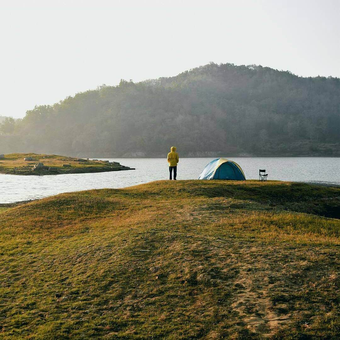 A person in a yellow jacket stands near a blue tent on the grassy shore of Sermo Reservoir, with a calm lake and hills in the background.