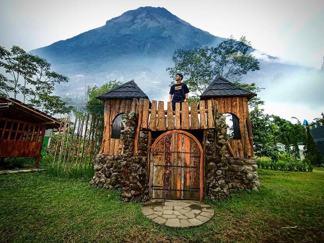 A visitor standing on a rustic wooden structure with an impressive view of Mount Merapi in the background at Bhumi Merapi Agrotourism.