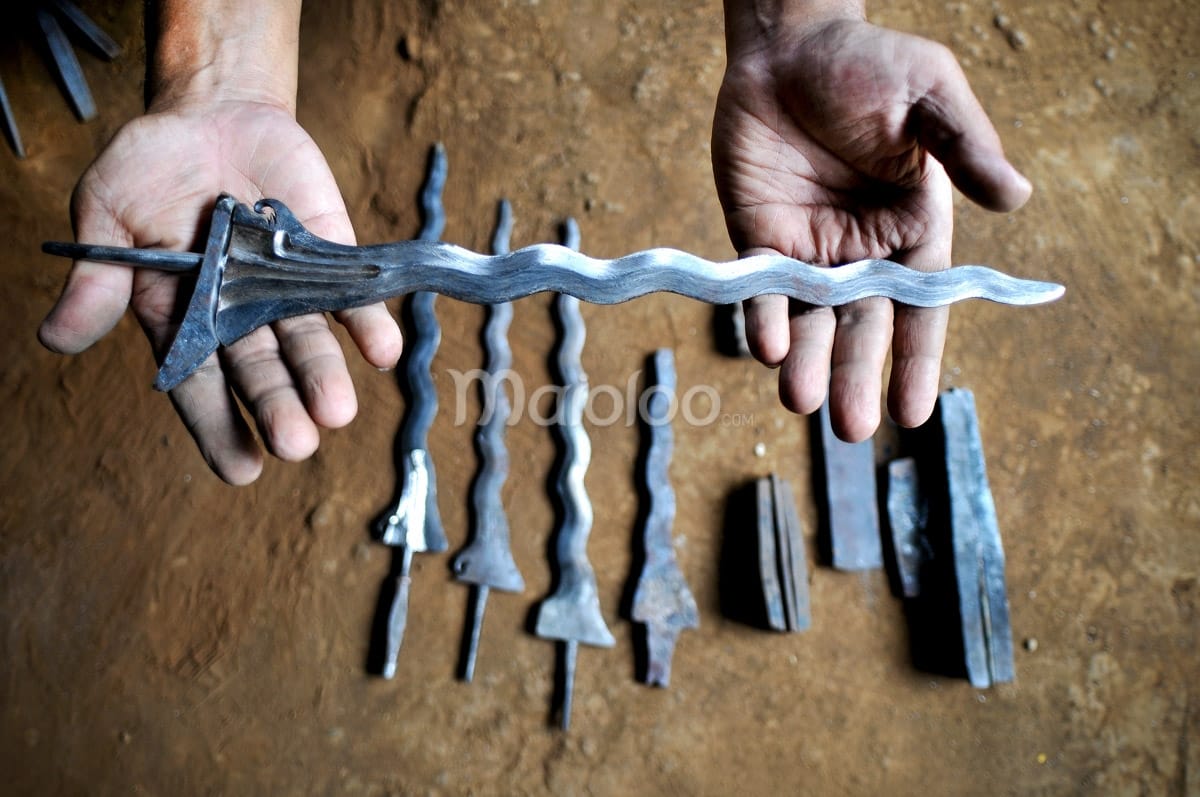 Close-up of a craftsman’s hands holding a nearly finished keris blade with a few other keris blades laid out on the ground.