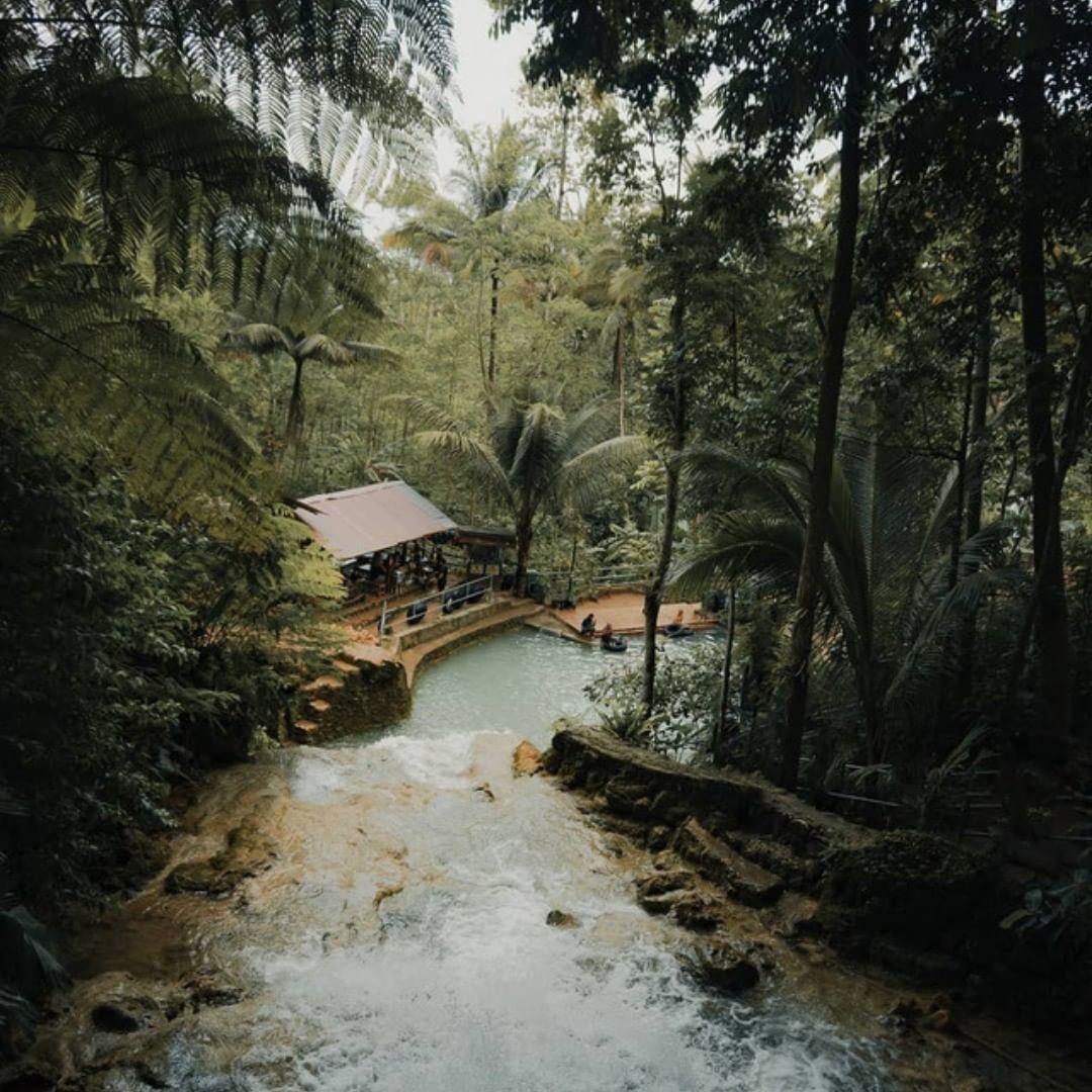 Aerial view of a lush green setting at Mudal River Park with a waterfall cascading into a pool surrounded by trees and a gazebo.