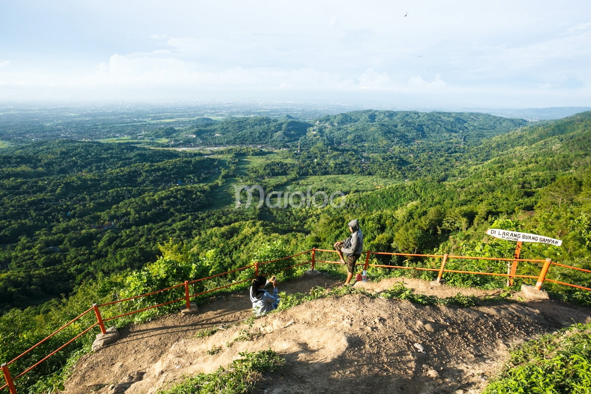 Two visitors enjoy the view from a vantage point at Becici Peak, with lush green forests and distant mountains visible.