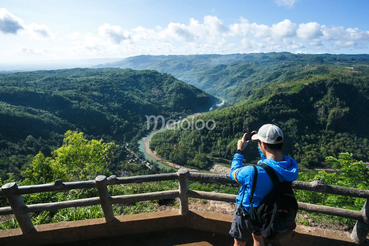 A person takes a photo from a viewing platform at Mangunan Fruit Garden, capturing the winding Oyo River and lush green hills below.