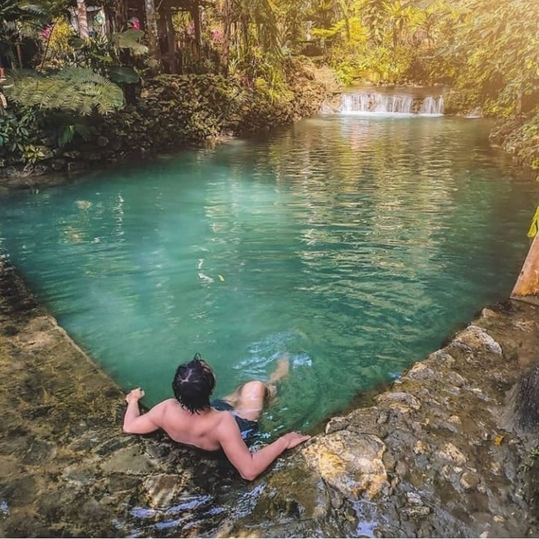 A man leans on the edge of a clear turquoise pool, enjoying the water with a small waterfall in the background at Mudal River Park.